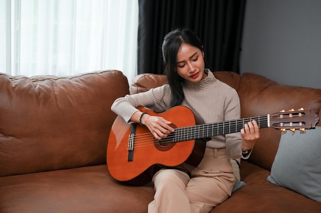 Talented and relaxed Asian female playing an acoustic guitar on her sofa in the living room