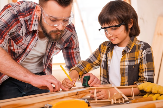 Talented workers. Concentrated young male carpenter teaching his son to work with wood in his workshop