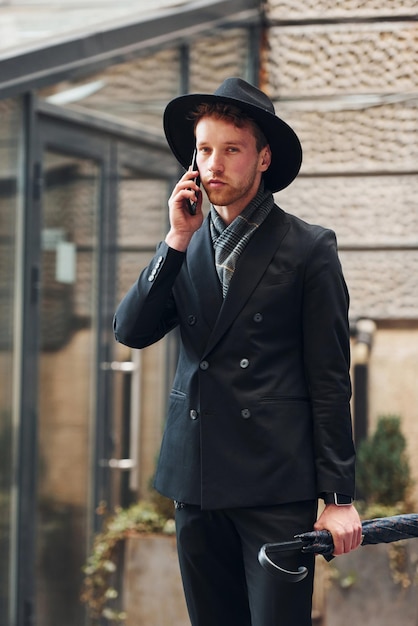 Talking by phone Elegant young man in black hat and clothes with umbrella in hands outdoors in the city