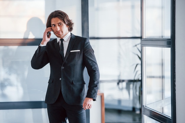 Talking by the phone in the office. Portrait of handsome young businessman in black suit and tie.