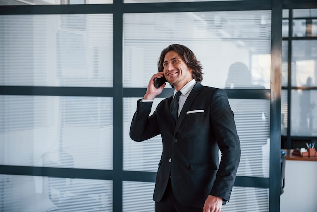 Talking by the phone in the office. Portrait of handsome young businessman in black suit and tie.