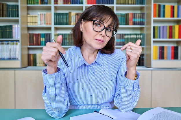 Talking female online teacher looking at virtual webcam while sitting in library office