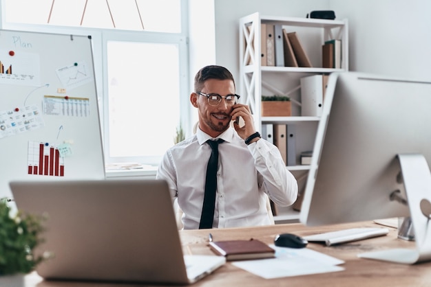 Talking with client. Handsome young man in formalwear talking on the phone and smiling while sitting in the office