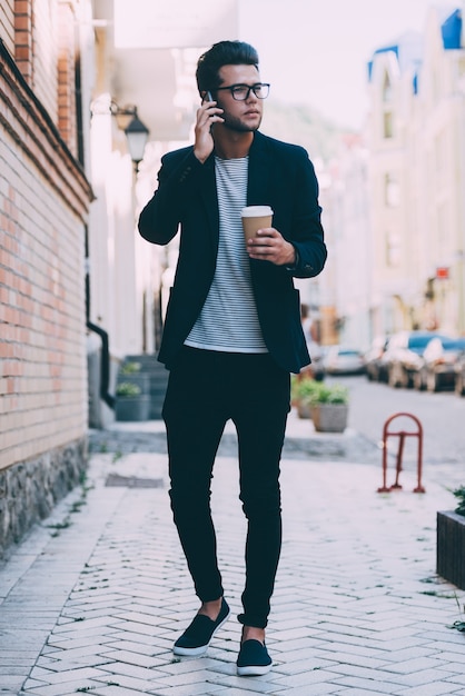 Talking with friend. Full length of handsome young man in smart casual wear carrying coffee cup and talking on mobile phone while walking along the street