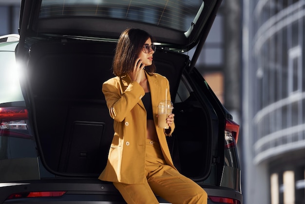 Talks by phone Young fashionable woman in burgundy colored coat at daytime with her car