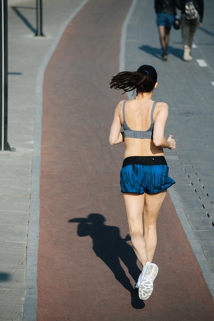 Tall athletic woman running away on the new track outdoors next to a walkway. On a sunny day under a clear blue sky. She is wearing grey top and mini shorts.