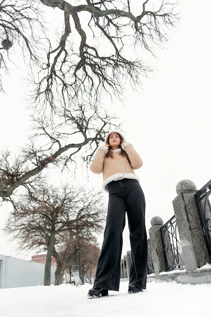 Tall young slender woman in black trousers and beige sheepskin coat in snowcovered park Stylish girl Vertical frame