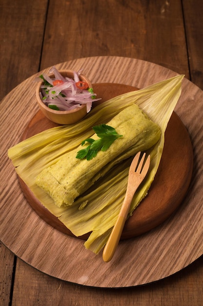 Photo tamales wrapped in banana leaves with a bowl of cilantro and a fork on a wooden plate.