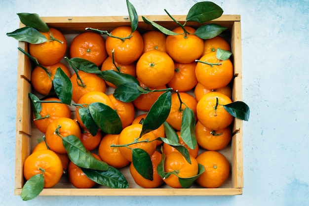 Tangerines oranges, clementines, citrus fruits with green leaves in a wooden box over light  with copyspace