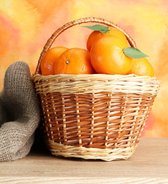 Tangerines with leaves in a beautiful basket on wooden table on orange background