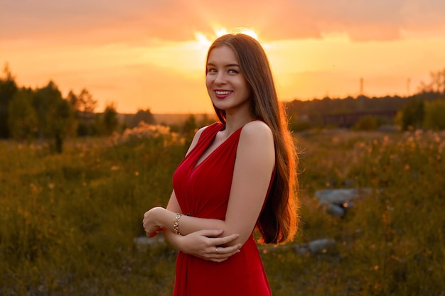 Tanned beautiful girl with long hair in a red dress in a field at sunset