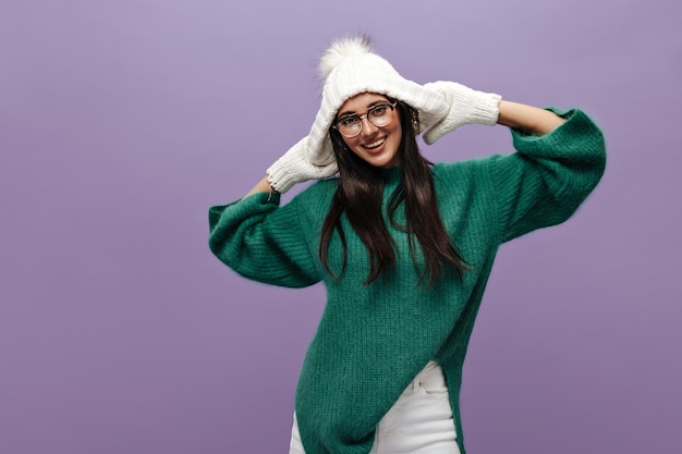 Tanned brunette woman in knitted white hat and gloves poses on purple background Cute girl in eyeglasses and green sweater smiles sincerely on isolated