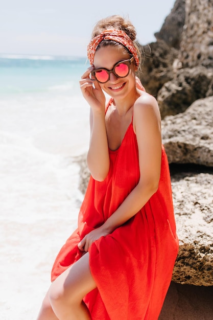 Tanned enchanting girl touching her glasses while sitting on rock Outdoor photo of pretty female model in red attire posing on ocean background