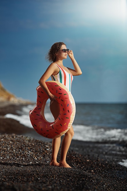 Tanned young woman in a swimsuit and sunglasses standing on a beach with an inflatable donut.