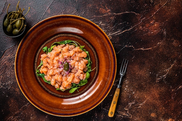 Tartar or tartare with salmon fish, red onion, arugula and capers in rustic plate. Dark background. Top View. Copy space.