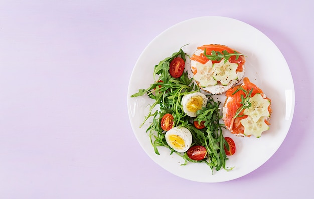 Tasty breakfast. Open sandwiches with salmon, cream cheese and rye bread in a white plate and salad with tomato, egg and arugula. Top view. Flat lay