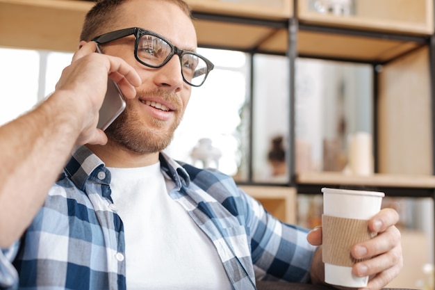 Tasty drink. Positive intelligent good looking man holding a plastic cup with coffee and enjoying his drink while having a conversation via cell phone