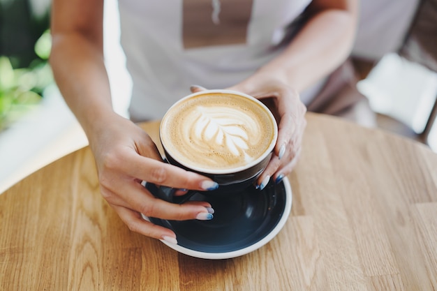 Tasty fresh cappuccino in cup on wooden table. Unrecognizable woman holding cup in hands.