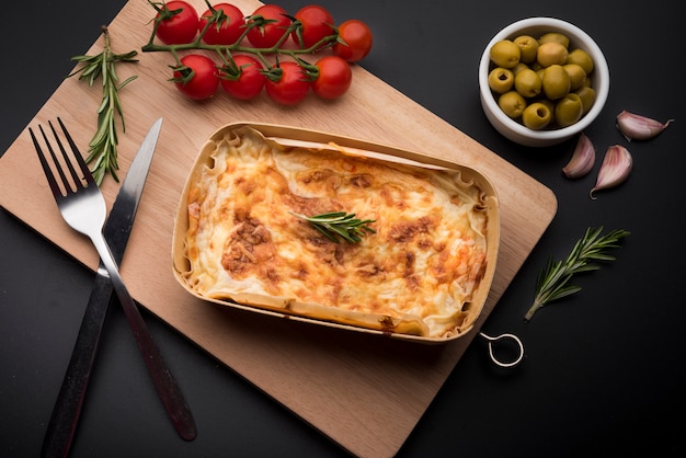 Tasty lasagna and ingredient on wooden cutting board over black surface