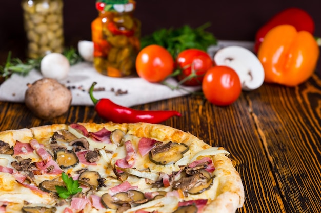 Tasty pizza with ham, onion and pickles on wooden table, tomatoes, mushrooms and other vegetables in the background