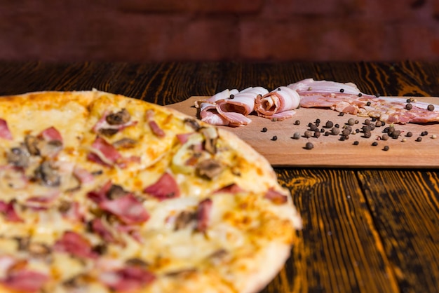 Tasty pizza with variety of toppings and cheese on wooden table, focus on cutting board with bacon and spices