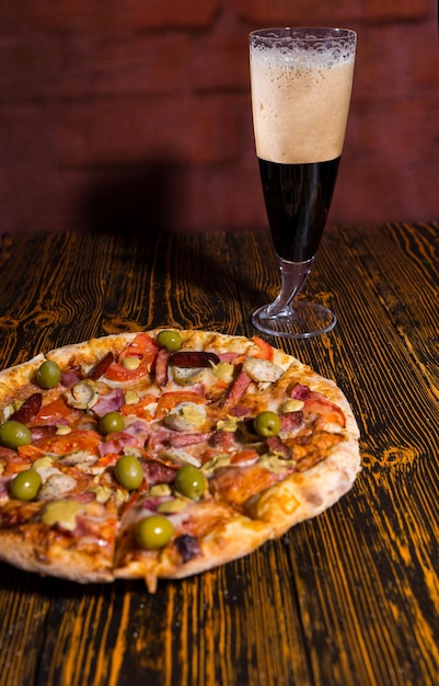 Tasty pizza with variety of toppings and cheese on wooden table near a glass of dark beer