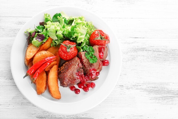 Photo tasty roasted meat with cranberry sauce and roasted vegetables on plate on color wooden background