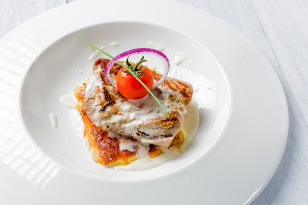 Tasty roasted pork pieces with cheese sauce, tomato and onion on white plate close up