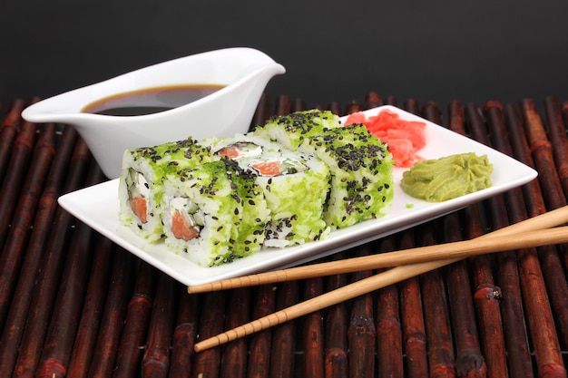 Tasty rolls served on white plate with chopsticks on bamboo mat on black background