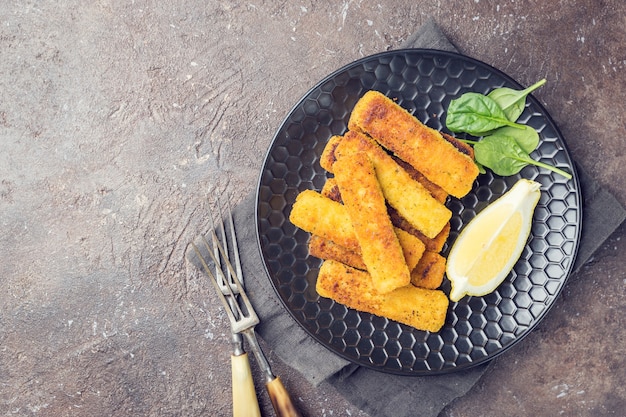 Tasty savory snack of crumbed fish fingers sticks served on a plate with lemon over dark stone