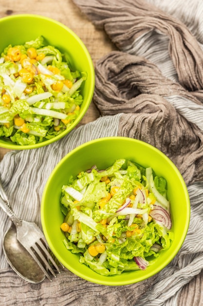 Tasty vegan salad from Chinese cabbage, sweet corn, purple onion, and dressing. Wooden background, cutlery, vintage napkin, rustic style, top view