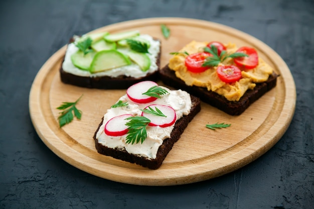 Tasty vegetarian rye bread toasts with cottage cheese, hummus, avocado, raddish and tomato on a wooden board