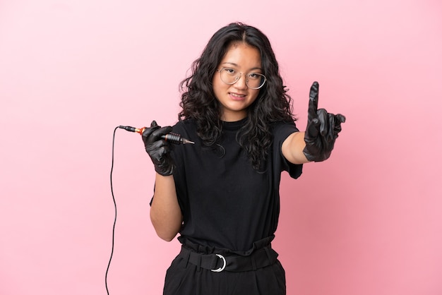 Tattoo artist asian woman isolated on pink background showing and lifting a finger