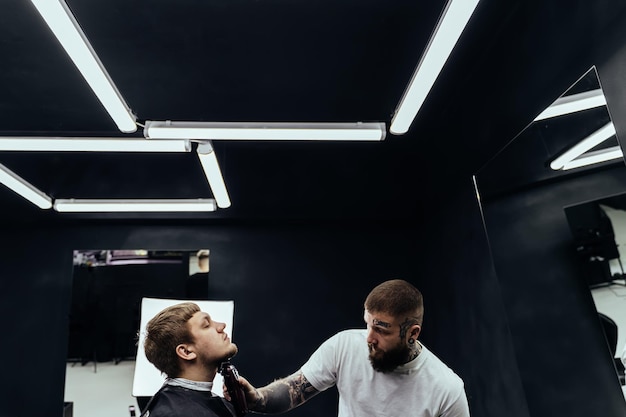 Tattooed Barber trimming bearded man with shaving machine in barbershop Hairstyling process Hairstylist cutting the beard of a bearded male