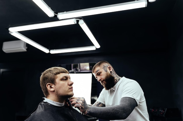 Tattooed Barber trimming bearded man with shaving machine in barbershop Hairstyling process Hairstylist cutting the beard of a bearded male