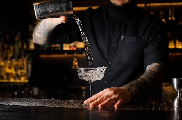 Tattooed bartender pouring an alcoholic drink from the steel shaker to the empty cocktail glass on the bar counter