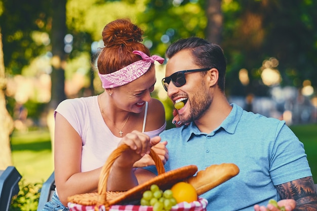 Tattooed bearded male and redhead female are having a picnic on a bench in a park.