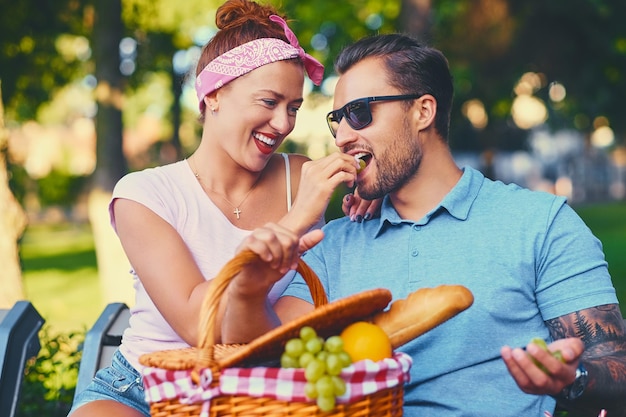 Tattooed bearded male and redhead female are having a picnic on a bench in a park.