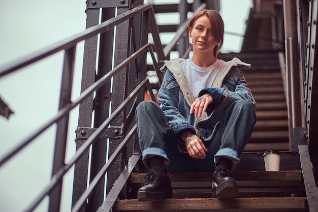 A tattooed redhead girl wearing trendy clothes sitting on stairs outside.