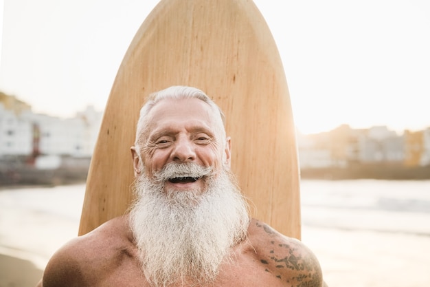 Tattooed senior surfer holding vintage surf board on the beach at sunset - Focus on face