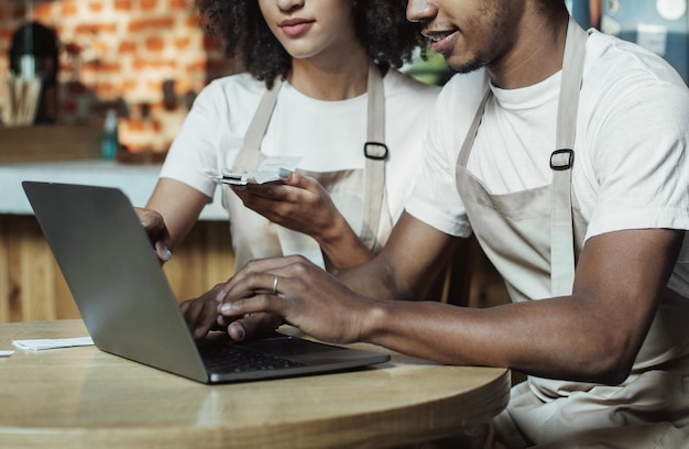 Taxes for family businesses during COVID-19 lockdown, bookkeeping, check bills in cafeteria. Busy millennial african american couple in aprons working on laptop in cafe interior, cropped, free space