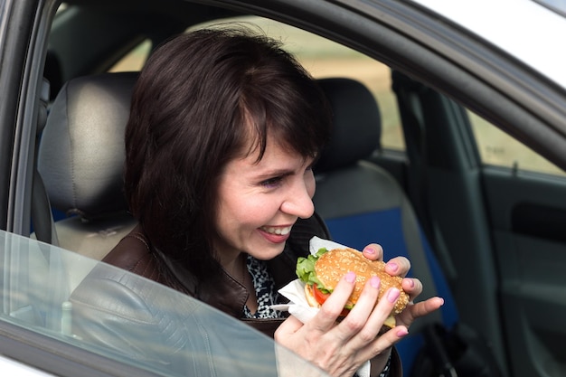 The taxi driver, laughing, in the car eats a juicy hamburger.