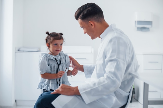 Teaches girl how to use stethoscope. Young pediatrician works with little female visitor in the clinic.