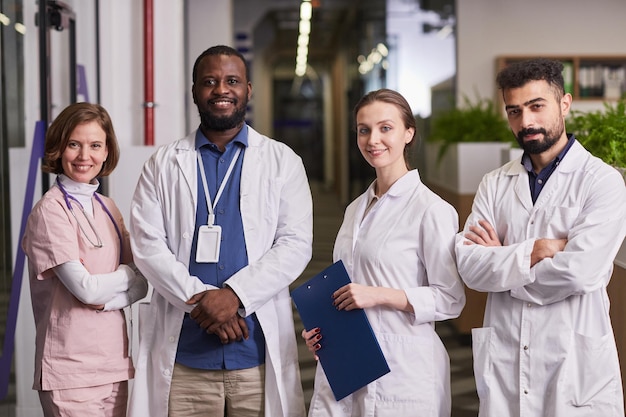 Team of happy young intercultural healthcare workers in uniform looking at camera while standing in row in corridor of modern clinics