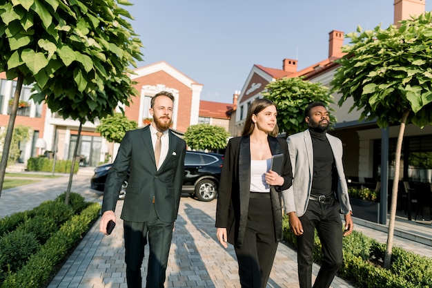 Team of multiethnical business people two men and woman in smart casual wear, walking outdoor on the business meeting. Woman holds digital tablet. Black car and modern buildings