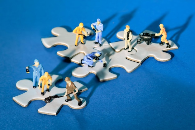 Team of tiny worker miniature figures on linked jigsaw puzzle pieces island on dark blue paper Dramatic light with long shadows