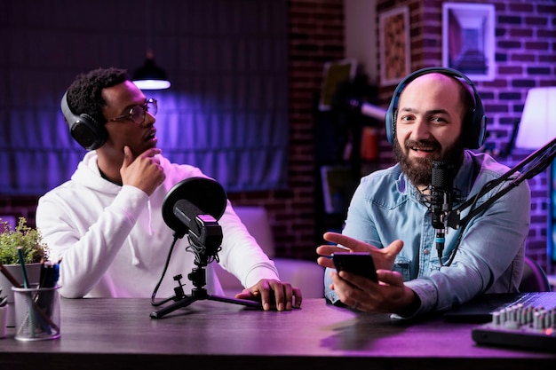 Team of vloggers filming podcast video show in modern studio, reading comments on smartphone. Male content creators recording vlog on camera and live streaming conversation on internet.