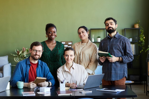 Team of young successful designers gathered by workplace looking at camera with smiles while working