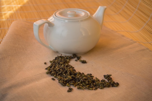 Teapot with green tea leaves on the napkin bamboo background