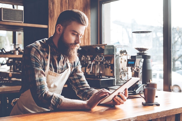 Technologies make business easier. Young bearded man in apron using his digital tablet while leaning to bar counter at cafe